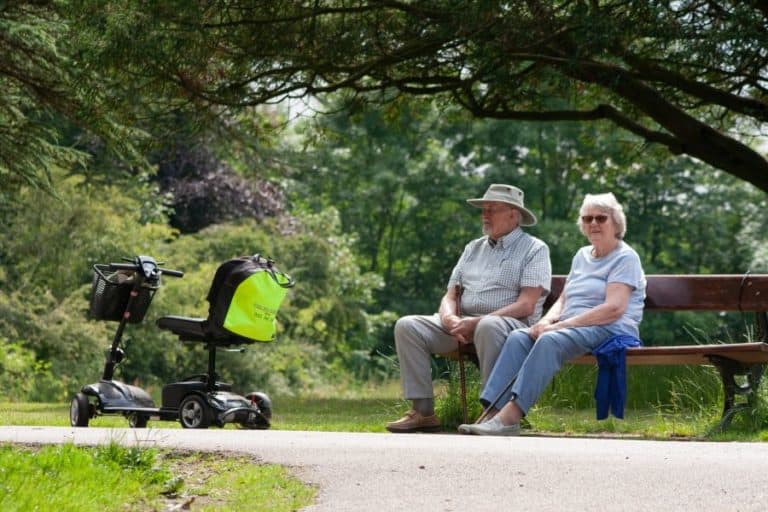 an elderly couple sits on a bench near an EV scooter powered by a charging station